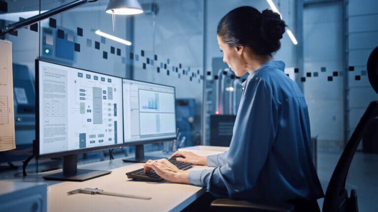 Mujer trabajando en oficina tecnológica con dos monitores, utilizando software especializado sobre diferentes sistemas operativos, en un entorno empresarial que depende de la infraestructura digital para análisis de datos y gestión de procesos.