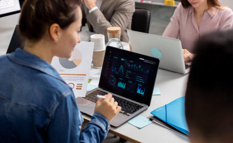 Mujer analizando datos en laptop mientras colegas revisan gráficos de ciencia de datos e inteligencia artificial en reunión corporativa.