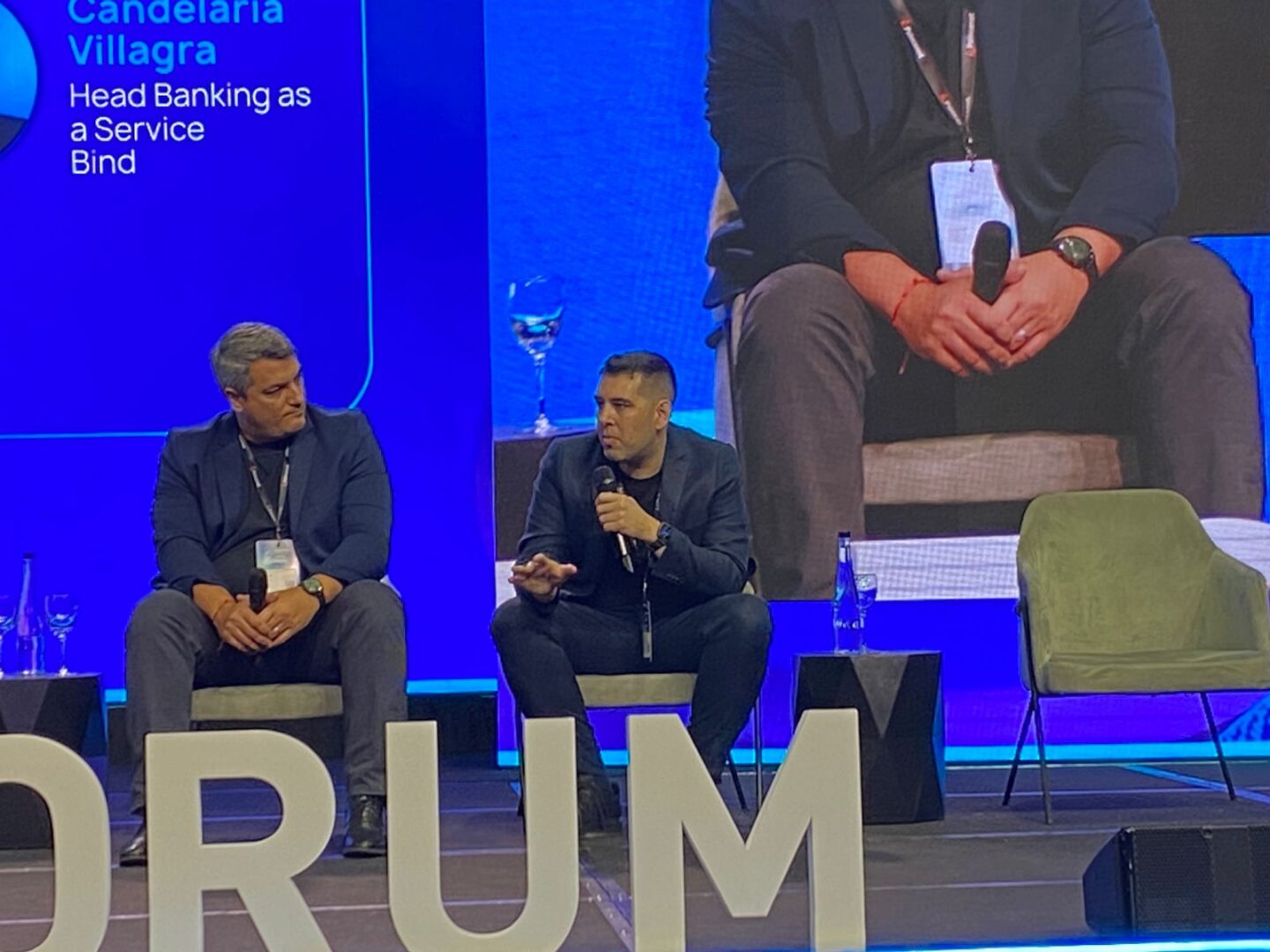 Dos panelistas sentados en un escenario del Argentina Fintech Forum dialogan sobre pagos digitales, con una pantalla azul de fondo que muestra datos del evento; según la información provista, el panel incluye a Cuello de Coelsa y a Sarria de Suga.
