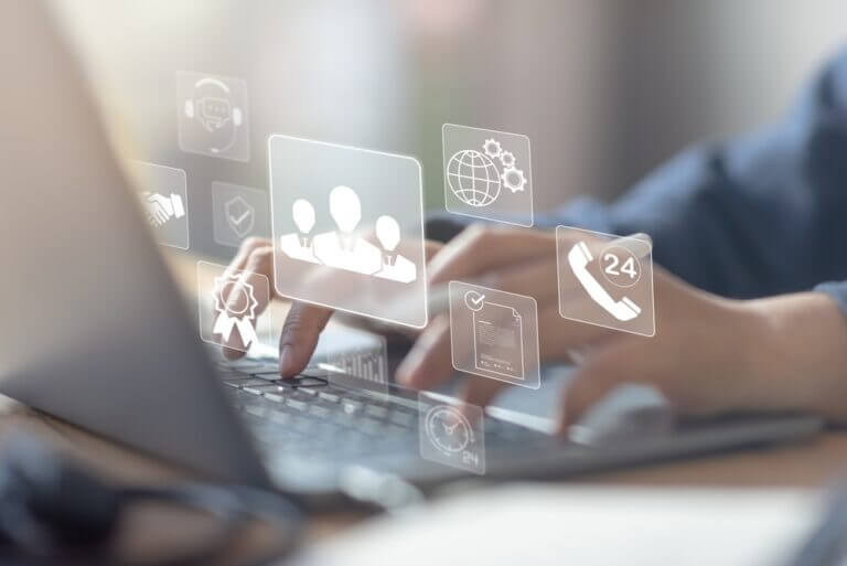 Persona escribiendo en un portátil mientras iconos de servicios digitales y soporte 24/7 se proyectan sobre la pantalla, simbolizando la gestión de recursos de TI en la empresa.