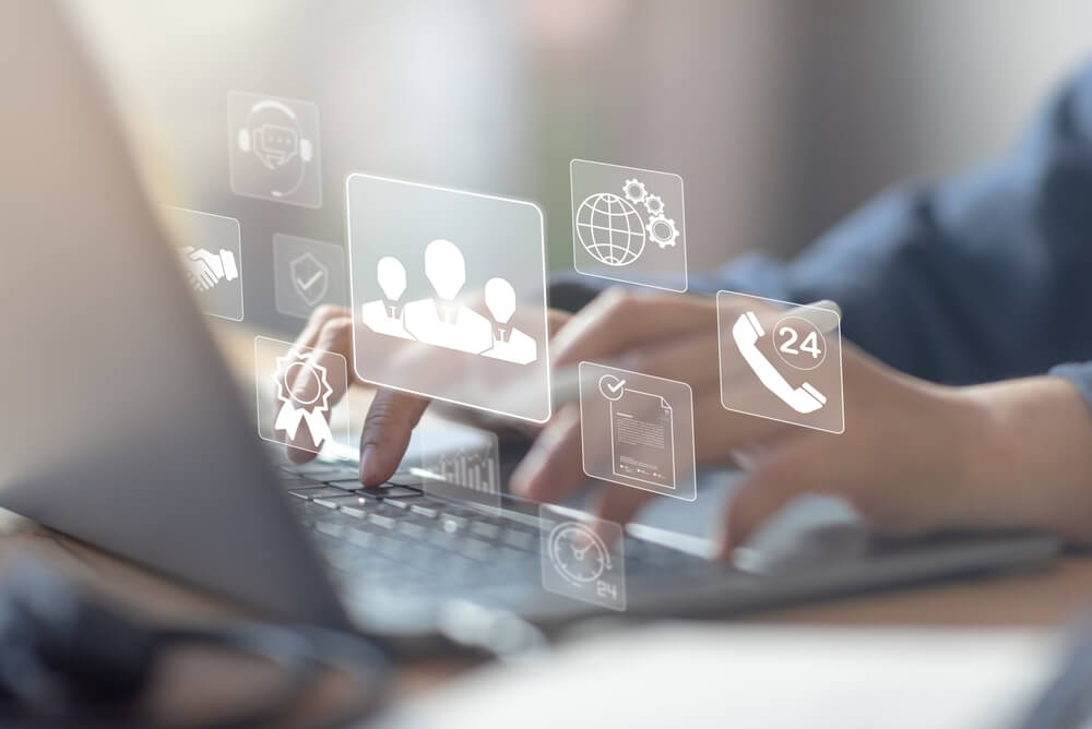 Persona escribiendo en un portátil mientras iconos de servicios digitales y soporte 24/7 se proyectan sobre la pantalla, simbolizando la gestión de recursos de TI en la empresa.