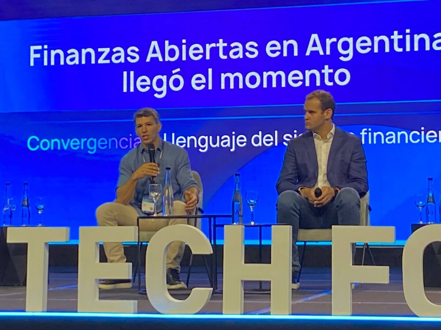 Dos integrantes del panel del Argentina Fintech Forum 2025 sentados en el escenario durante una exposición sobre Open Finance, con una pantalla azul que destaca el tema central del encuentro.