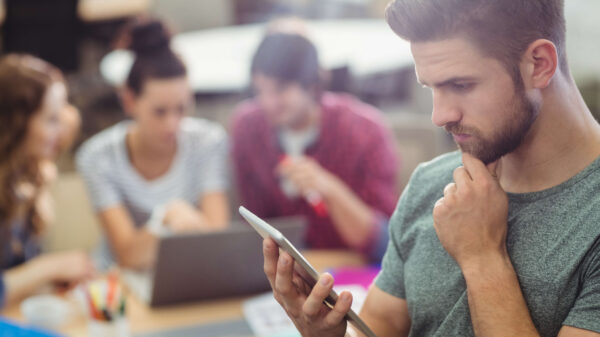 Alumno usando el celular para interactuar con un chatbot educativo, con otros estudiantes utilizando computadoras en el fondo. Representa el uso de herramientas de IA para resolver consultas y apoyar el aprendizaje.