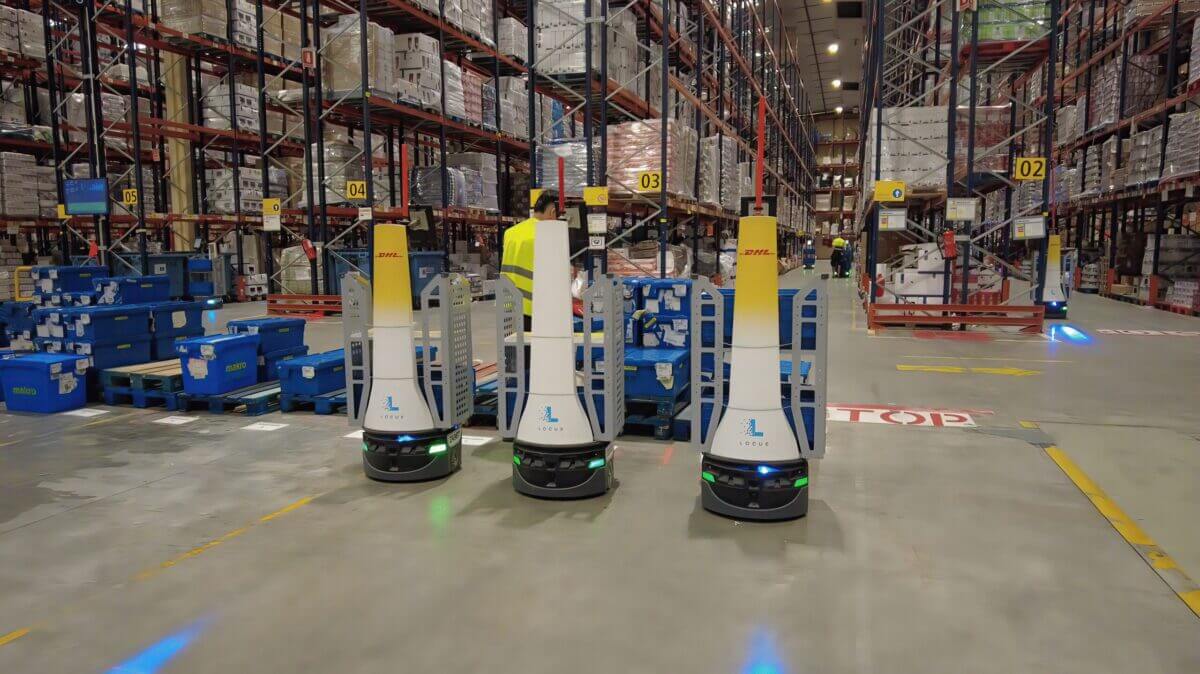 Tres robots móviles autónomos AMR de Locus Robotics operan en un depósito de DHL junto a estanterías, estaciones de picking y contenedores logísticos.