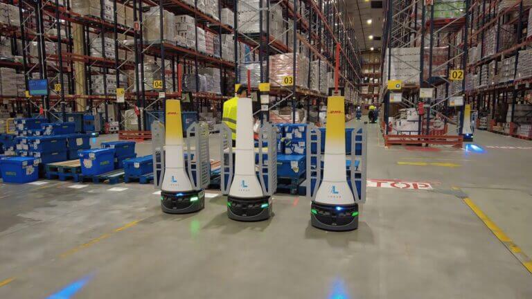 Tres robots móviles autónomos AMR de Locus Robotics operan en un depósito de DHL junto a estanterías, estaciones de picking y contenedores logísticos.