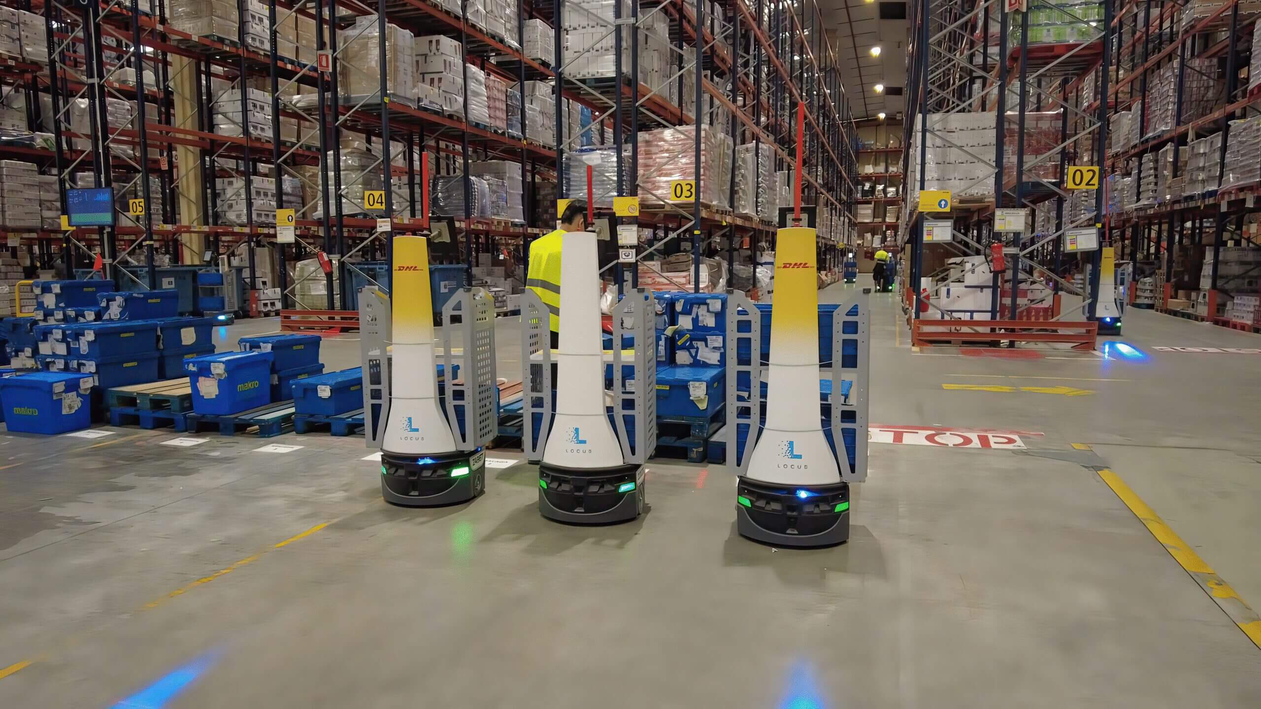 Tres robots móviles autónomos AMR de Locus Robotics operan en un depósito de DHL junto a estanterías, estaciones de picking y contenedores logísticos.