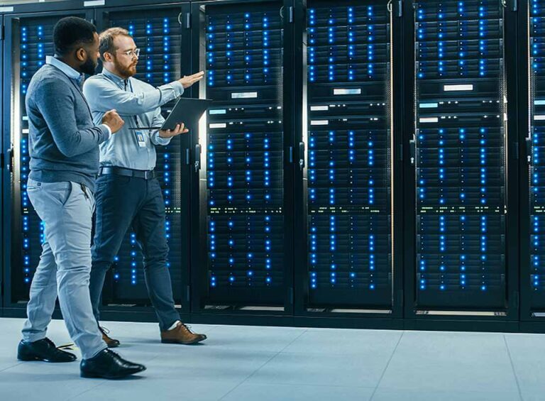 Dos técnicos caminan con una laptop entre racks de servidores, en una imagen sobre gestión de data centers e infraestructura crítica.