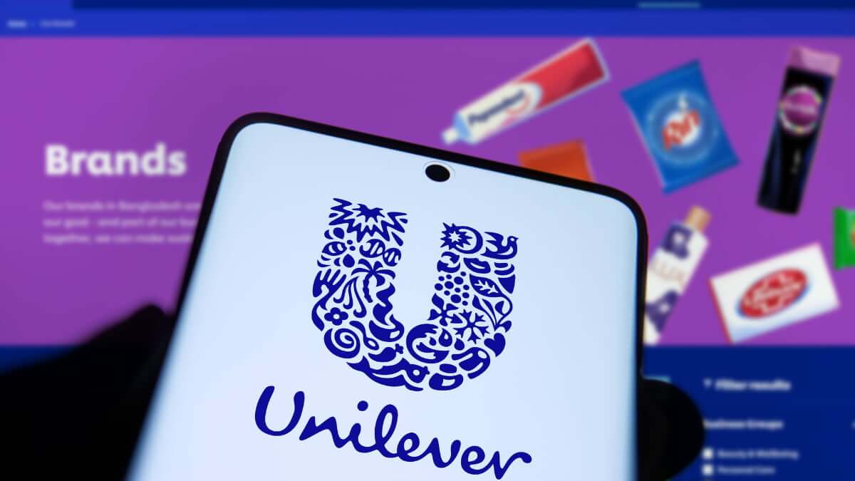Logo de Unilever en la pantalla de un celular frente a imágenes de marcas de consumo, en una nota sobre IA, GenAI y automatización industrial.