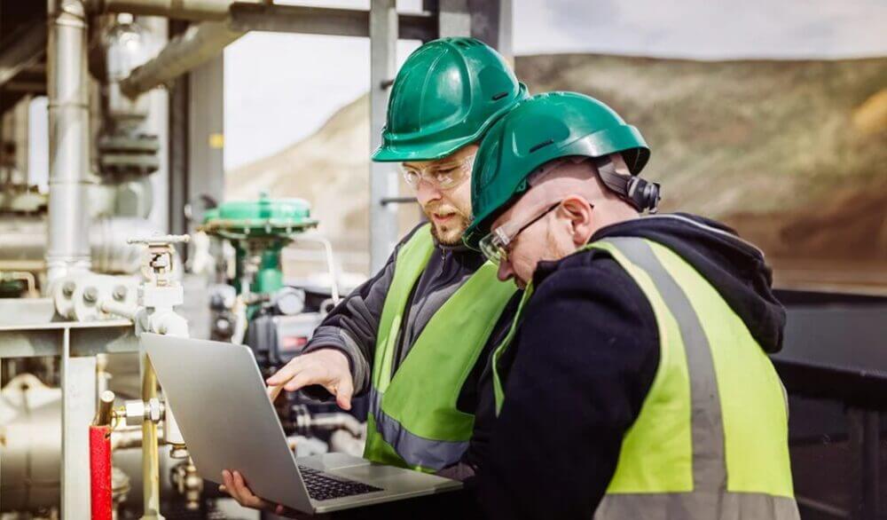 Dos técnicos con casco revisan una laptop junto a equipos industriales, en una escena asociada a automatización en refinerías y APC.