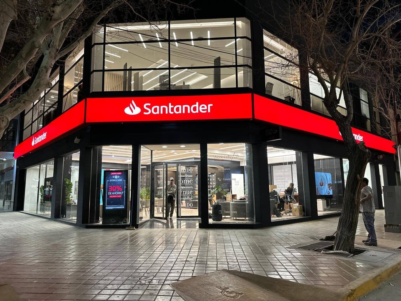 Fachada iluminada de una sucursal Santander Work Café con cartelería roja, vidrieras amplias y espacios de atención en el interior.