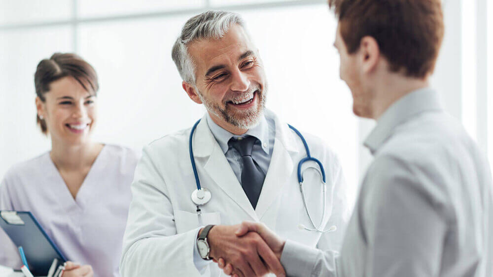 Médico saluda a un paciente junto a una profesional de salud, en una escena vinculada a Patient 360 y a la integración de datos para mejorar la atención.