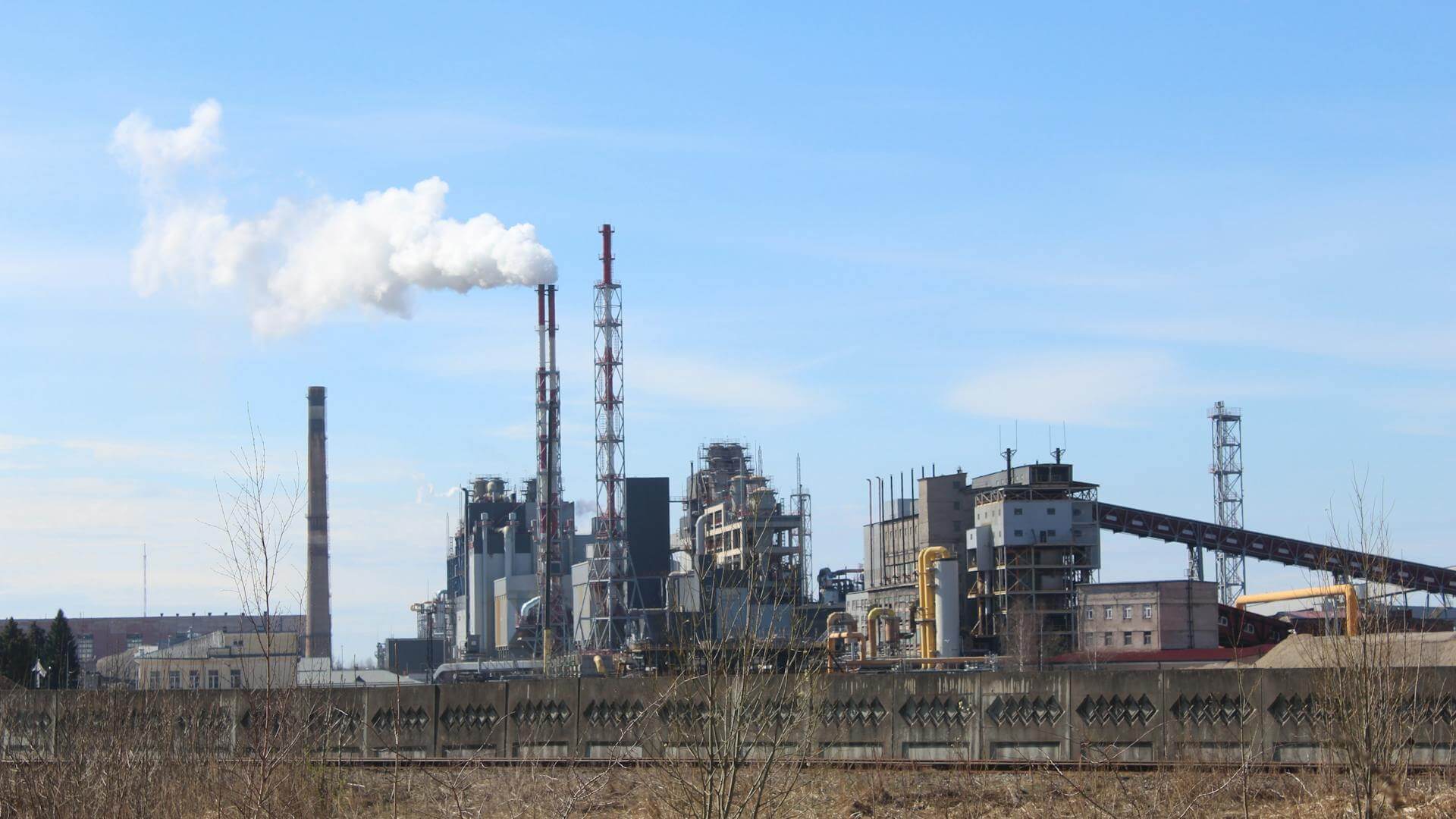 Vista exterior de una planta industrial con chimeneas, estructuras productivas y humo saliendo de una torre.