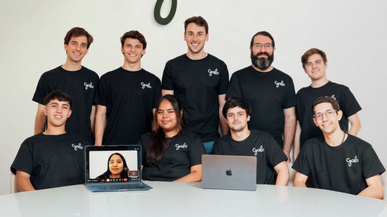 Equipo de Galo AI posa en una oficina con remeras de la empresa, dos notebooks sobre la mesa y una integrante conectada por videollamada.
