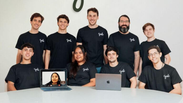 Equipo de Galo AI posa en una oficina con remeras de la empresa, dos notebooks sobre la mesa y una integrante conectada por videollamada.