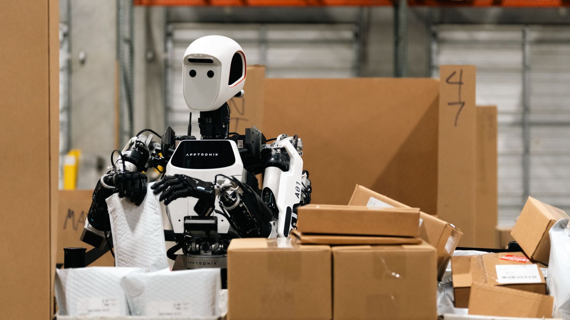 Robot humanoide Apollo de Apptronik en Robot Park, entre cajas de cartón dentro de un área de pruebas logísticas.