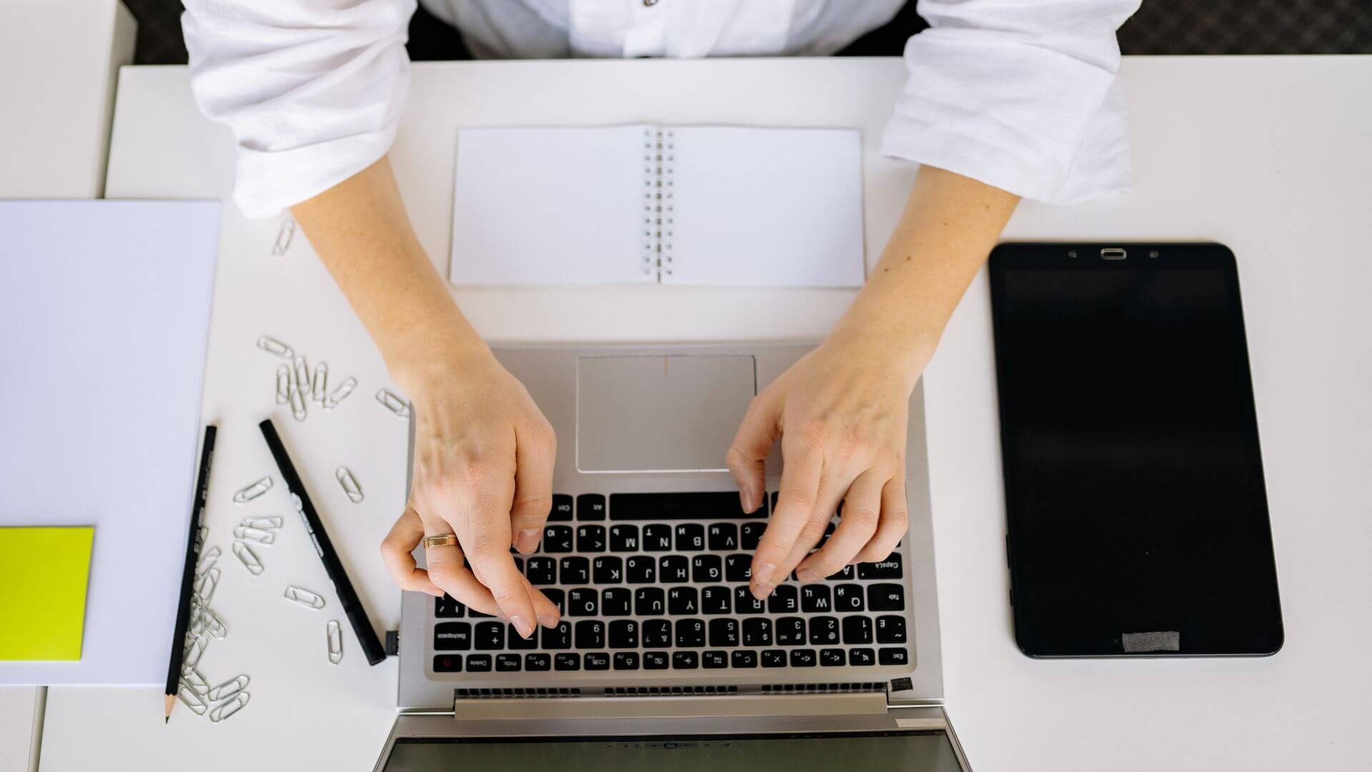 Vista superior de manos sobre una notebook, con cuaderno, tablet y útiles de escritorio, escena vinculada al primer empleo.