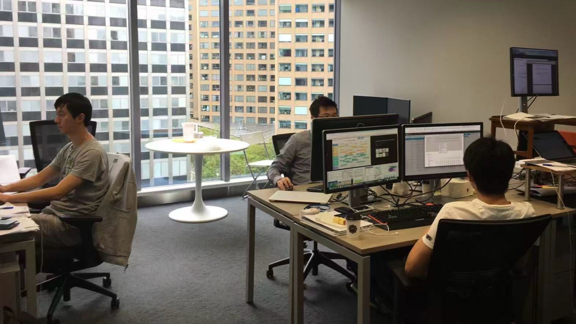 Tres empleados trabajan frente a computadoras en las primeras oficinas de Airwallex, con edificios urbanos visibles a través de los ventanales.