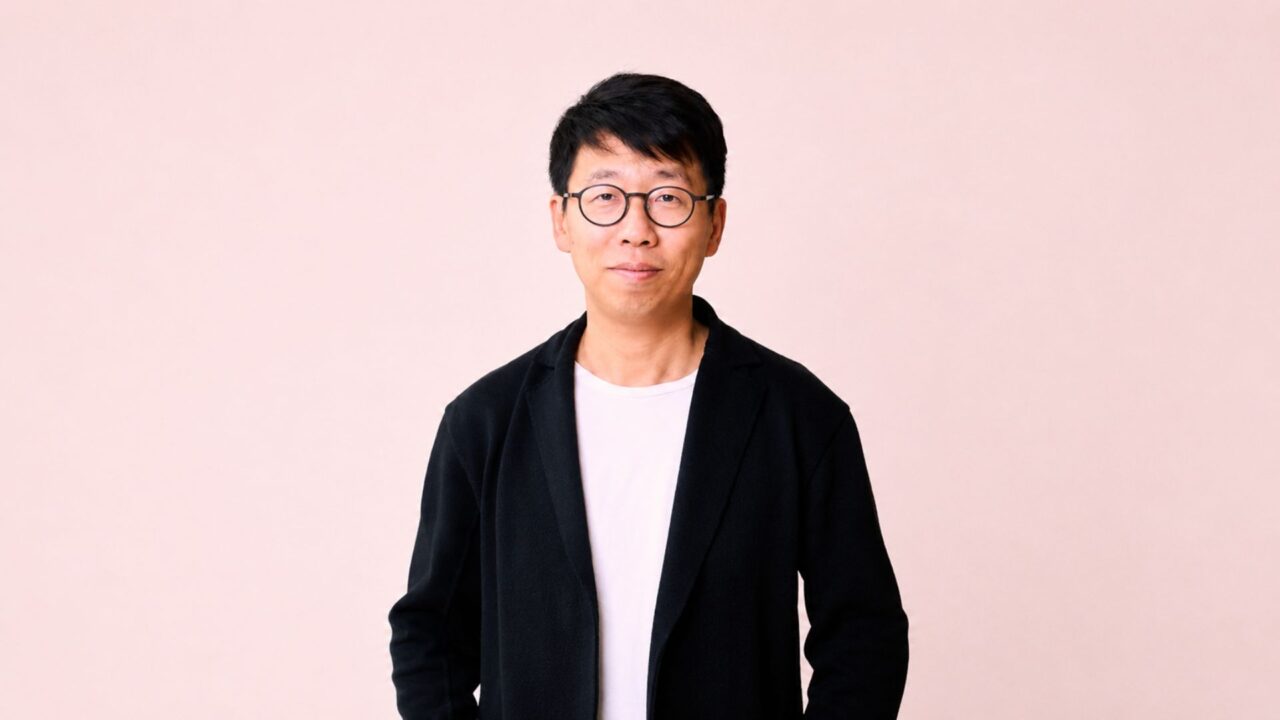 Retrato de un ejecutivo con anteojos y saco negro sobre fondo rosa, en una imagen corporativa de Airwallex.