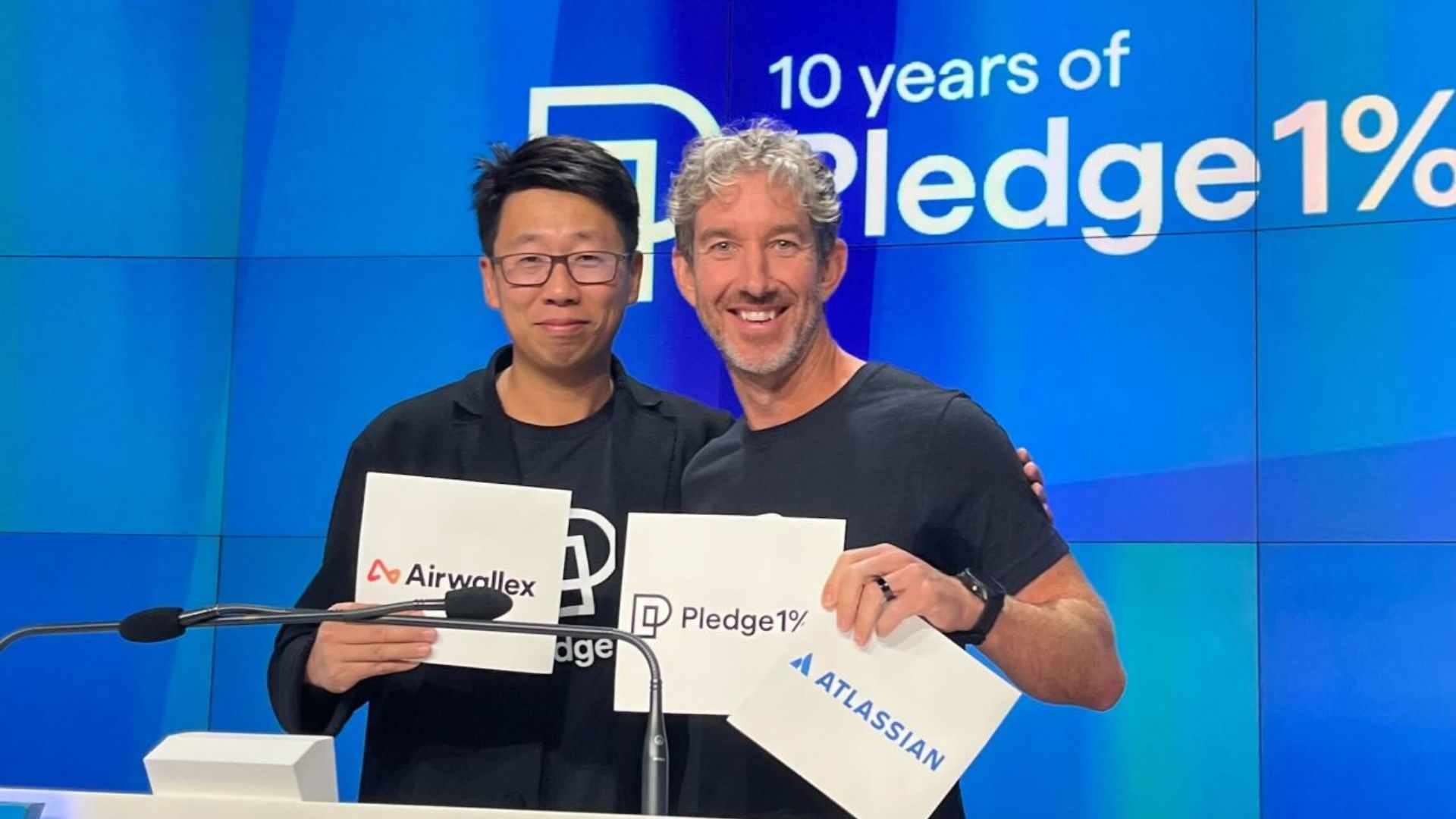 Dos ejecutivos posan con carteles de Airwallex, Pledge 1% y Atlassian frente a una pantalla azul durante un encuentro corporativo.