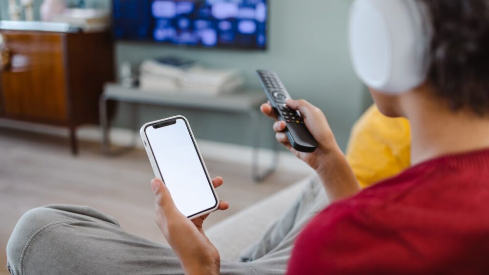 Usuario con auriculares sostiene un teléfono y un control remoto frente a una plataforma de streaming abierta en el televisor.