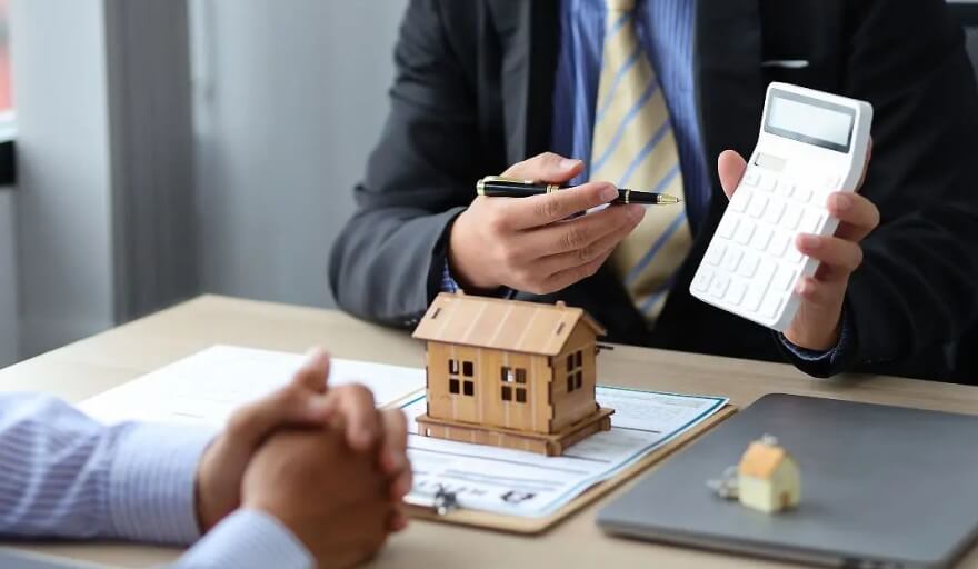 Asesor financiero muestra una calculadora junto a una maqueta de vivienda durante una evaluación crediticia.