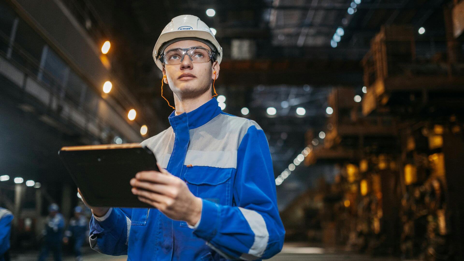 Ingeniero de campo con casco, protección auditiva y una tablet dentro de una planta industrial.