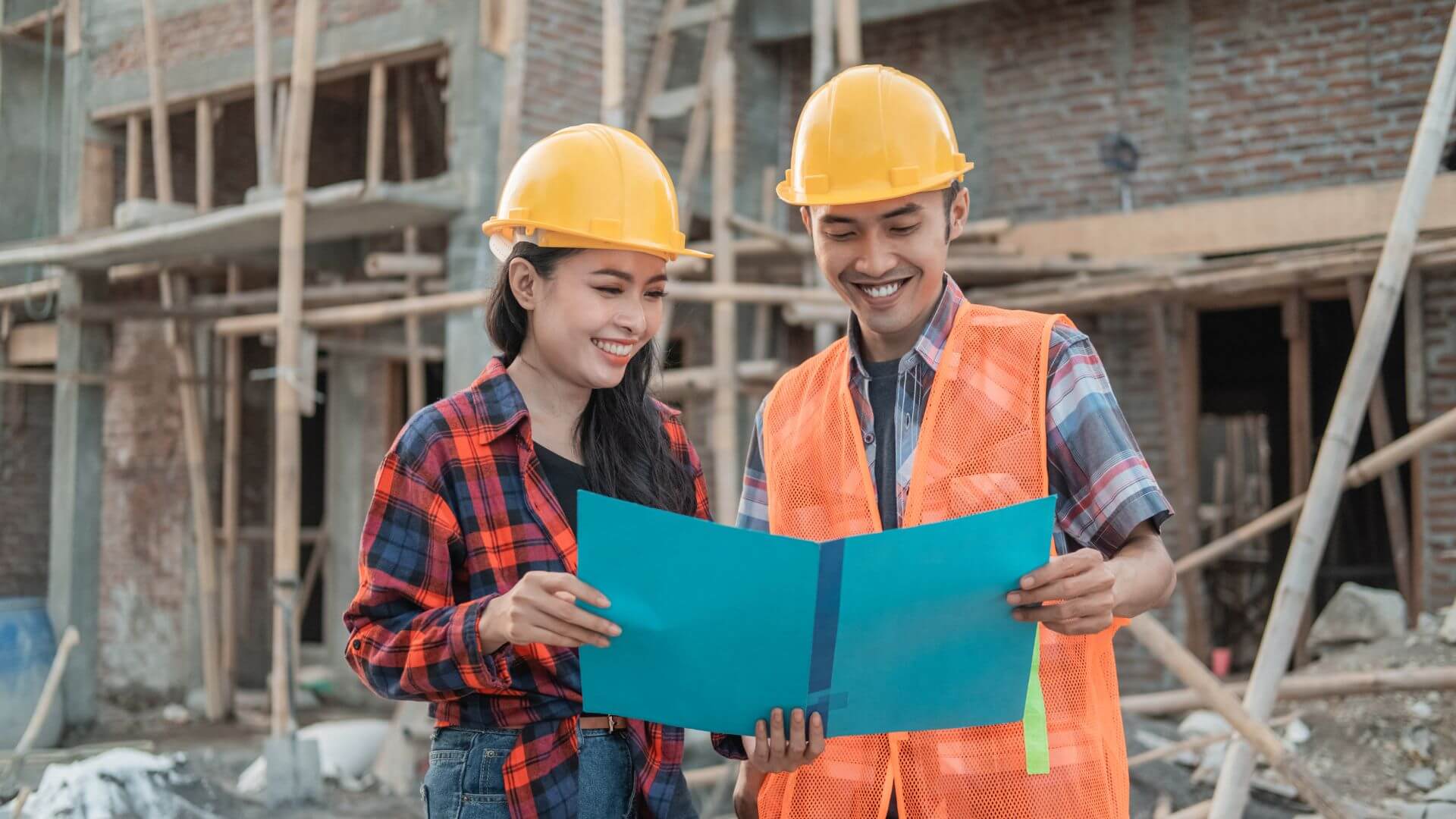 Dos ingenieros civiles revisan planos frente a una obra en construcción.