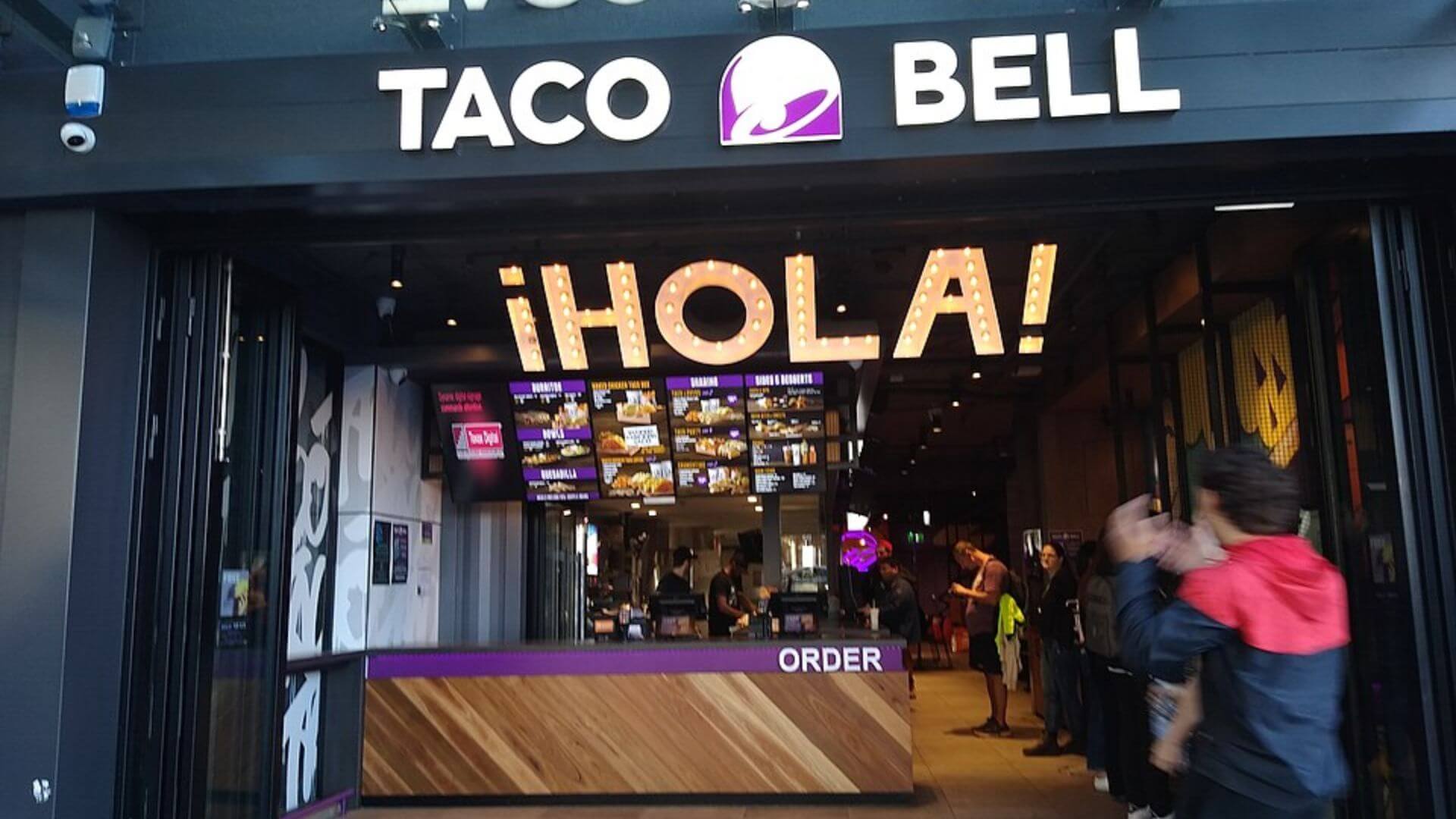 Interior de un restaurante Taco Bell con mostrador de pedidos, cartelería digital y clientes cerca de la entrada.