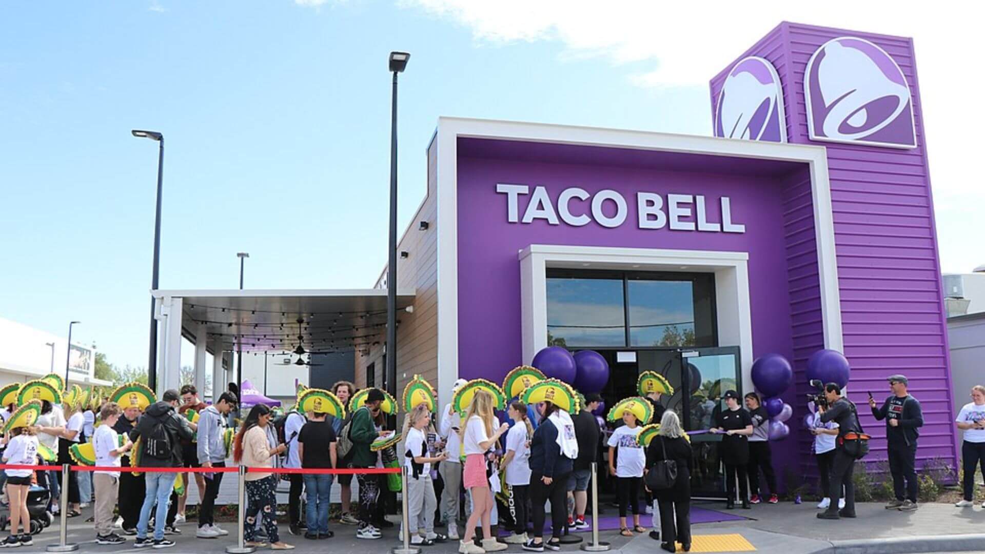 Fachada violeta de un restaurante Taco Bell con clientes reunidos frente a la entrada durante un evento.