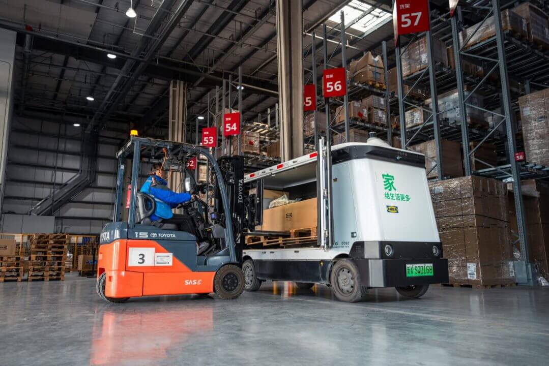 Vehículo autónomo de IKEA junto a una carretilla elevadora durante una operación de carga en un depósito de China.